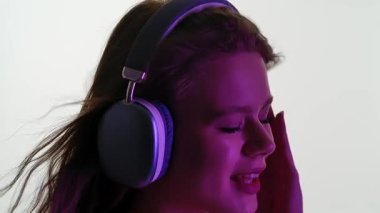 Genç bir kadın, canlı mor bir ışığın altında ritme dalmış, büyük kulaklıklarla müzik dinliyor. Yavaş çekim