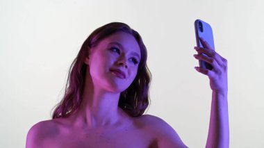 Genç bir kadın akıllı telefonuyla renkli bir ışık altında selfie çekerken gülümsüyor..