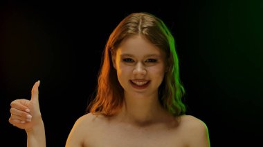 Bir kadın, siyah bir fona başparmak işareti yaparken gülümsüyor. Işıklandırma canlı ve pozitif bir atmosfer yaratıyor..