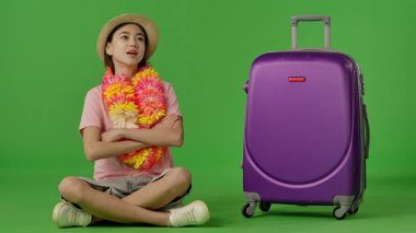 Çiçek kolyeli genç bir kız mor bavulun yanında oturuyor, kameraya bakıyor ve gülümsüyor. Krom anahtar yeşil ekran yeşil ekran stüdyo arka plan modelinde izole edilmiş. Yavaş çekim