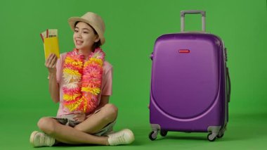 Çiçekli genç kız mor bavulun yanında oturuyor. Pasaport ve uçak biletlerini gösteriyor. Krom anahtar yeşil ekran stüdyo arka planda izole edilmiş Yavaş çekim