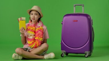 Çiçekli çiçek desenli genç kız mor bavulun yanında oturuyor. Pasaportu, uçak biletlerini ve jestlerini gösteriyor. Krom anahtar yeşil ekran yeşil ekran stüdyo arka plan modelinde izole edilmiş. Yavaş çekim