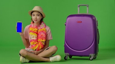 Çiçekli genç kız mor bavulun yanında oturuyor, mavi ekranlı akıllı telefonu işaret ediyor, kopyalama alanı. Krom anahtar yeşil ekran yeşil ekran stüdyo arka plan modelinde izole edilmiş. Yavaş çekim