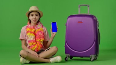 Çiçekli genç kız mor bavulun yanında oturuyor, mavi ekranlı akıllı telefonu işaret ediyor, kopyalama alanı. Krom anahtar yeşil ekran yeşil ekran stüdyo arka plan modelinde izole edilmiş. Yavaş çekim