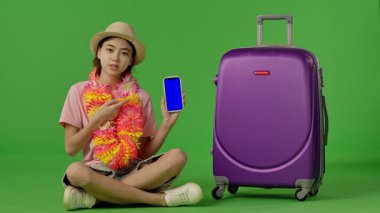 Çiçekli genç kız mor bavulun yanında oturuyor, mavi ekranlı akıllı telefonu işaret ediyor, kopyalama alanı. Krom anahtar yeşil ekran yeşil ekran stüdyo arka plan modelinde izole edilmiş. Yavaş çekim