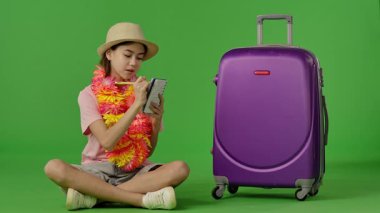 Çiçek kolyeli genç bir kız mor bavulun yanında oturuyor ve not defterine yazıyor. Krom anahtar yeşil ekran yeşil ekran stüdyo arka plan modelinde izole edilmiş. Yavaş çekim