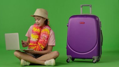 Çiçek kolyeli genç kız mor bavulun yanında oturuyor ve dizüstü bilgisayarda yazıyor. Krom anahtar yeşil ekran yeşil ekran stüdyo arka plan modelinde izole edilmiş. Yavaş çekim
