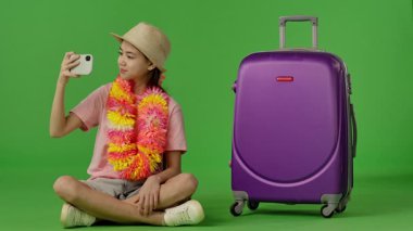 Çiçekli Ley giyen genç bir kız mor bavulun yanında oturmuş akıllı telefondan selfie çekiyor. Krom anahtar yeşil ekran yeşil ekran stüdyo arka plan modelinde izole edilmiş. Yavaş çekim
