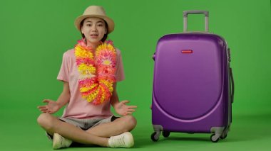 Çiçek kolyeli genç bir kız mor bavulun yanında oturuyor, kameraya bakıyor ve gülümsüyor. Krom anahtar yeşil ekran yeşil ekran stüdyo arka plan modelinde izole edilmiş. Yavaş çekim