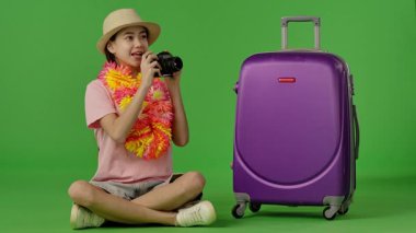Çiçekli Ley giyen genç kız mor bavulun yanına oturur ve retro kamerayla fotoğraf çeker. Krom anahtar yeşil ekran yeşil ekran stüdyo arka plan modelinde izole edilmiş. Yavaş çekim