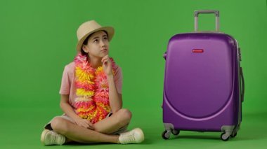 Çiçek kolyeli genç bir kız mor bavulun yanında oturuyor ve bir şey düşünüyor. Krom anahtar yeşil ekran yeşil ekran stüdyo arka plan modelinde izole edilmiş. Yavaş çekim