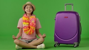 Çiçek kolyeli genç bir kız mor bavulun yanında oturuyor, kameraya bakıyor ve gülümsüyor. Krom anahtar yeşil ekran yeşil ekran stüdyo arka plan modelinde izole edilmiş. Yavaş çekim
