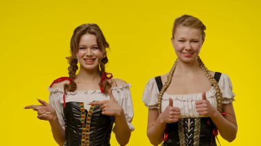 Geleneksel Bavyera elbiseli iki gülümseyen kadın, parlak sarı bir arka plana karşı poz veriyorlar..