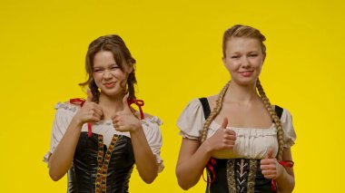 Geleneksel Bavyera elbiseli iki gülümseyen kadın, parlak sarı bir arka plana karşı poz veriyorlar..