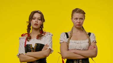 Saçları örgülü geleneksel kostümler giyen iki kadın, parlak sarı bir arkaplanda duruyorlar..