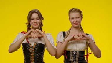 Geleneksel Bavyera giysileri giyen iki kadın sarı arka planda kalp sembolleri yapıyorlar..