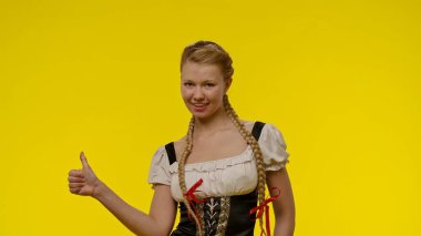 Saçları örgülü geleneksel kostümlü gülümseyen genç bir kadın sarı bir arka plana başparmak atar..