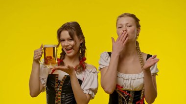Geleneksel kostümlü iki kadın, canlı sarı bir arka planda bira eşliğinde eğlenceli bir anın tadını çıkarıyorlar..