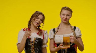 Geleneksel Bavyera kostümlü iki kadın, ellerinde bira bardağı, parlak sarı bir arka plana gülümsüyorlar..