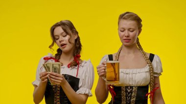 Geleneksel Bavyera elbiseli iki kadın, ellerinde bir yığın para ve parlak sarı arka planda bir bira bardağı tutarken görülüyor..