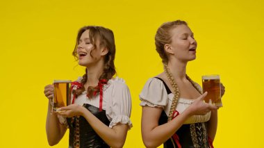 Geleneksel kostümlü iki kadın, canlı sarı bir arka planda bira fincanlarını birlikte kutlayarak eğleniyorlar..