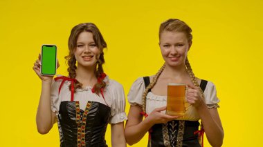 Geleneksel Bavyera kostümü giymiş iki kadın ellerinde yeşil ekranlı bir akıllı telefon ve parlak sarı bir fonda bira bardağı tutuyorlar..
