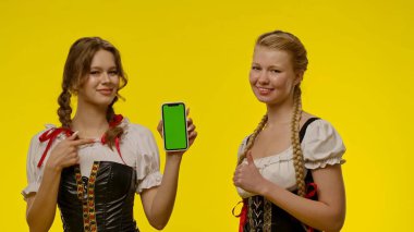 Geleneksel Bavyera kostümü giymiş iki kadın ellerinde yeşil ekranlı bir akıllı telefon, parlak sarı bir arka plana karşı stein.