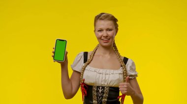 Gülümseyen genç bir kadın yeşil ekran ile akıllı bir telefon tutuyor, başparmağını kaldırıyor, parlak sarı bir arka fona dayanıyor..