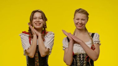 Geleneksel kostümler içinde gülümseyen ve poz veren iki kadın, canlı sarı bir arka planda..