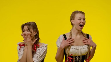 Geleneksel Dirndl elbiseli iki kadın, canlı sarı bir arkaplanda heyecan ifade ediyorlar. Giysileri arasında örgülü saç şekilleri ve dekoratif fiyonklar var. Şenlikli ve canlı bir atmosfer yaratıyorlar..