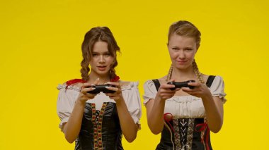 Geleneksel elbiseler giyen iki genç kadın, sarı bir zemine karşı büyük bir hevesle video oyunları oynuyor..