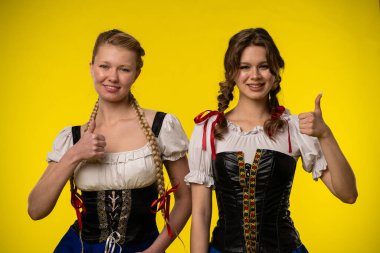 Geleneksel Bavyera elbiseli iki gülümseyen kadın, parlak sarı bir arka plana karşı poz veriyorlar..