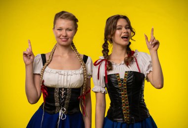 Atkuyruklu ve geleneksel kıyafetli iki genç kadın, parlak sarı bir zemin karşısında heyecanlanıyor..