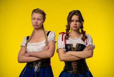 Saçları örgülü geleneksel kostümler giyen iki kadın, parlak sarı bir arkaplanda duruyorlar..