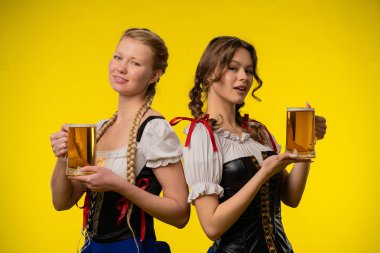 Geleneksel kıyafetler giyen iki kadın bira bardakları tutuyor, parlak sarı bir arka plan önünde gülümsüyorlar..