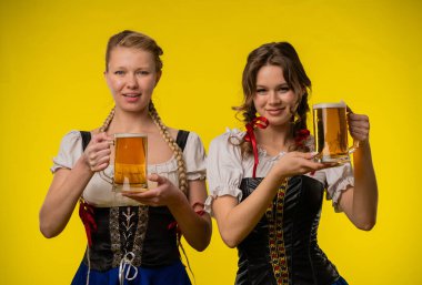 Geleneksel kıyafetler giyen iki kadın bira bardakları tutuyor, parlak sarı bir arka plan önünde gülümsüyorlar..