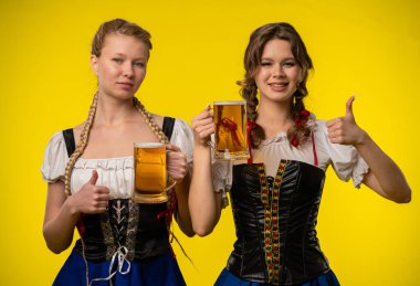Geleneksel Bavyera elbiseli iki gülümseyen kadın, parlak sarı bir arka plana karşı poz veriyorlar..
