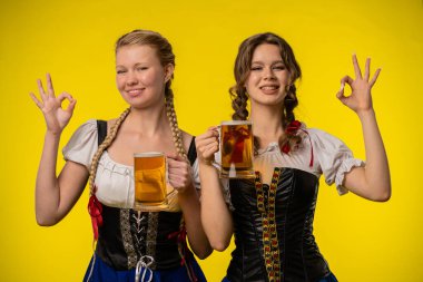 Geleneksel elbiseler giyen iki kadın, parlak sarı bir zemine karşı iyi jestler yapıyor, özgüven ve tarz yayıyorlar..