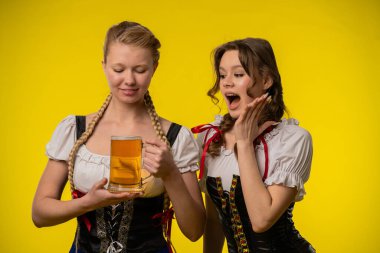 Geleneksel kıyafetli iki kadın bir bira bardağını tutarak, parlak sarı bir arka plana karşı neşeli ve neşeli bir atmosfer yaratıyorlar..