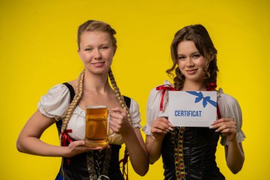 Geleneksel giyinmiş iki kadın kutlama yaparken görülüyor, ellerinde bir sertifika ve bir bira bardağı parlak sarı bir fonda.