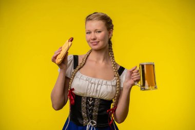 Geleneksel giyinmiş genç bir kadın, canlı sarı bir arka planda büyük bir sosis ve bir kupa biranın keyfini çıkarıyor..