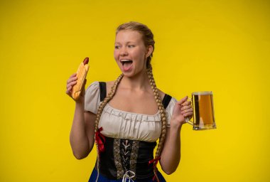 Geleneksel giyinmiş genç bir kadın, canlı sarı bir arka planda büyük bir sosis ve bir kupa biranın keyfini çıkarıyor..