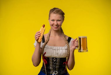 Geleneksel giyinmiş genç bir kadın, canlı sarı bir arka planda büyük bir sosis ve bir kupa biranın keyfini çıkarıyor..