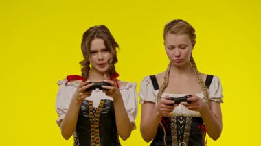Geleneksel elbiseler giyen iki genç kadın sarı bir zemine karşı hevesle video oyunları oynuyor. Yavaş çekim