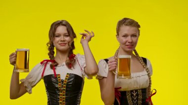 Geleneksel kıyafetler giyen iki kadın bira bardakları tutuyor, canlı sarı bir arkaplan önünde gülümsüyor ve dans ediyorlar. Yavaş çekim