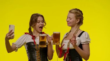 Geleneksel kostümler giyen iki kadın, bira fincanlarının keyfini çıkarıyorlar. Parlak sarı bir arka plana karşı selfie çekiyorlar. Neşeli bir anı yakalıyorlar..
