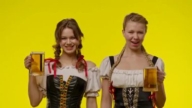 Geleneksel kostümlü iki kadın, canlı sarı bir arka planda bira fincanlarını birlikte kutlayarak eğleniyorlar..