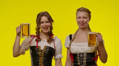 Geleneksel kıyafetler giyen iki kadın bira bardakları tutuyor, parlak sarı bir arka plan önünde gülümsüyorlar..