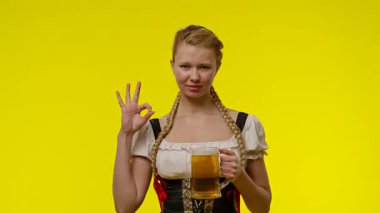 Geleneksel Bavyera elbisesi giymiş neşeli genç bir kadın elinde bir bardak bira tutuyor ve canlı sarı bir arka plana karşı mutlu poz veriyor..