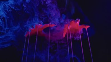 Bir çift el, canlı duman ve dramatik ışıklandırmadan oluşan ruhani bir manzarada ipleri manipüle eder. Yavaş çekim